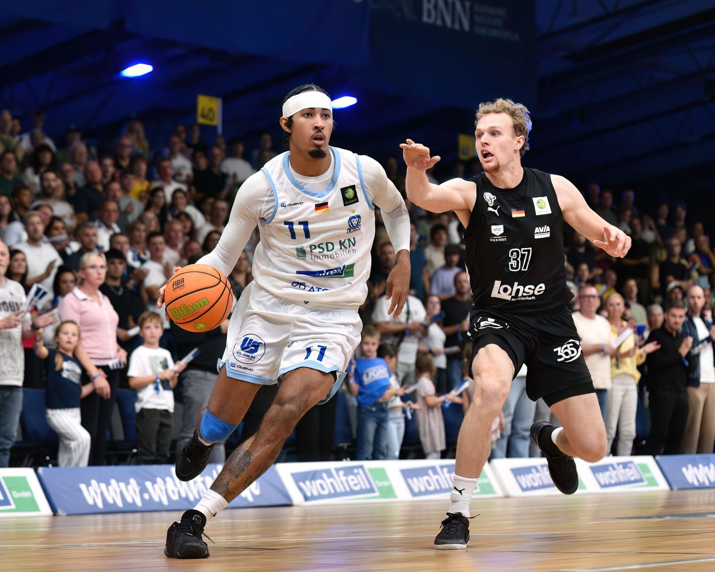 Jason George im Dribbling, in den Rängen dahinter Kinder und Familien