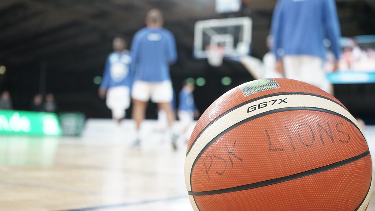 Spielball mit der Aufschrift PSK LIONS, dahinter die Mannschaft beim Aufwärmen
